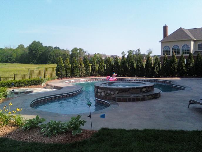 swimming pool with round stone covered spill over hot tub. Stamped concrete patio with step down fire pit. Pool features bluestone pool coping, cantilevered, sun shelf, deep-end bench, and colored led lights in Northville, Mi
